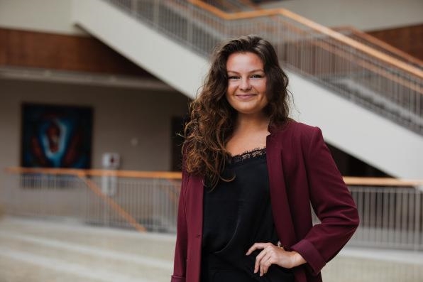 [ai] A woman with curly hair wearing a burgundy blazer and a black top, standing confidently in a modern building with a staircase in the background.